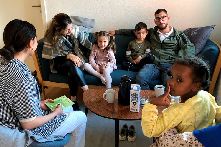Tosprogede børn og deres familier er samlet omkring højtlæsning i en sofa. En ældre pige sidder med en bog i skødet. || Hoejtlaesning Web Voksne Hoved 900X600