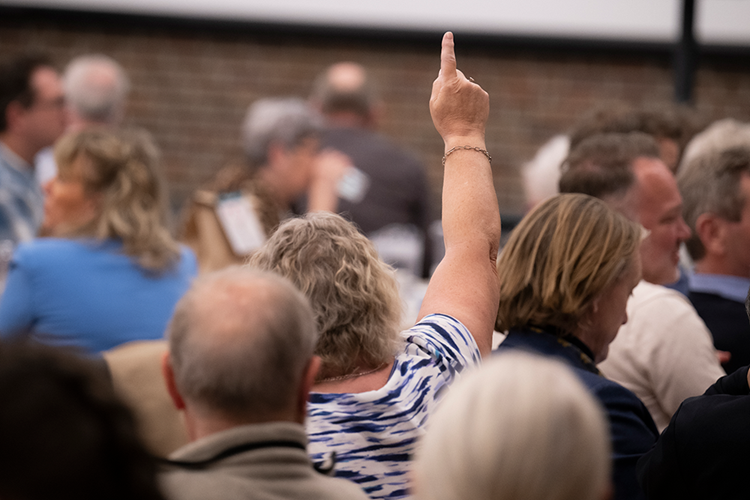 En stor gruppe af mennesker samlet i en stor sal - en kvinde med ryggen til rækker fingeren i vejret. || 2023Landskonf JL 0073 Forside Valg